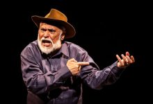 Cena do espetáculo “O Julgamento de Zé Bebelo”, com ator caracterizado como sertanejo gesticulando em palco escuro.