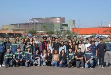Grupo de trabalhadores e colaboradores posa para foto em frente a complexo industrial com galpões e chaminés ao fundo, em dia ensolarado.