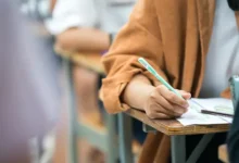 Pessoa sentada em carteira escolar escreve em folha de prova com lápis durante concurso público, enquanto outros candidatos aparecem desfocados ao fundo.