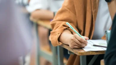 Pessoa sentada em carteira escolar escreve em folha de prova com lápis durante concurso público, enquanto outros candidatos aparecem desfocados ao fundo.