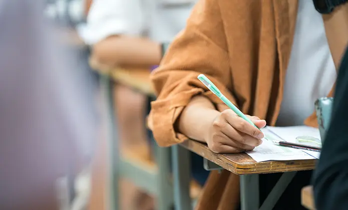 Pessoa sentada em carteira escolar escreve em folha de prova com lápis durante concurso público, enquanto outros candidatos aparecem desfocados ao fundo.