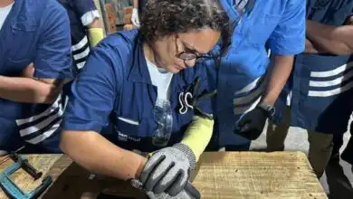 Pessoa utilizando equipamentos de proteção individual realiza atividade prática em bancada durante treinamento técnico, acompanhada por outros participantes em ambiente de capacitação profissional.