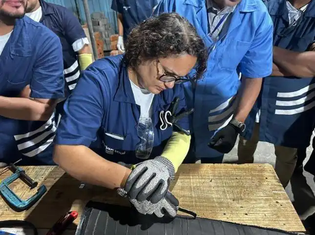 Pessoa utilizando equipamentos de proteção individual realiza atividade prática em bancada durante treinamento técnico, acompanhada por outros participantes em ambiente de capacitação profissional.