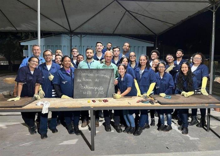 Grupo de participantes de curso de capacitação profissional posa para foto em ambiente de treinamento, usando uniformes e equipamentos de proteção, com bancada de trabalho ao fundo.