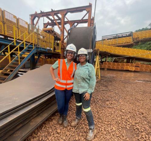 Duas trabalhadoras usam capacete e equipamentos de segurança em área industrial, posando em frente a estruturas metálicas e esteiras de operação.