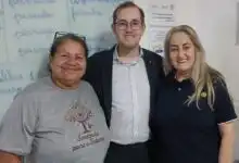 Três pessoas posam sorrindo dentro de uma sala de aula. Ao centro, o presidente do Rotary de Itaguaí, Josephj Piñeiro, de terno e óculos; à esquerda, Adeilda, presidente da ONG, que veste uma camiseta com a identificação da entidade “Semeando para o Futuro”; à direita, Eliane, mãe de Joseph e também rotariana, vestindo uma camisa polo com símbolo do Rotary. Ao fundo a lousa doada com palavras escritas, indicando ambiente educacional.