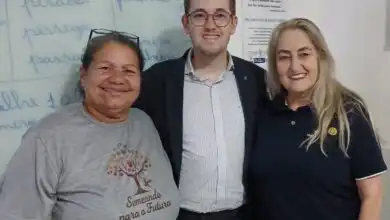 Três pessoas posam sorrindo dentro de uma sala de aula. Ao centro, o presidente do Rotary de Itaguaí, Josephj Piñeiro, de terno e óculos; à esquerda, Adeilda, presidente da ONG, que veste uma camiseta com a identificação da entidade “Semeando para o Futuro”; à direita, Eliane, mãe de Joseph e também rotariana, vestindo uma camisa polo com símbolo do Rotary. Ao fundo a lousa doada com palavras escritas, indicando ambiente educacional.