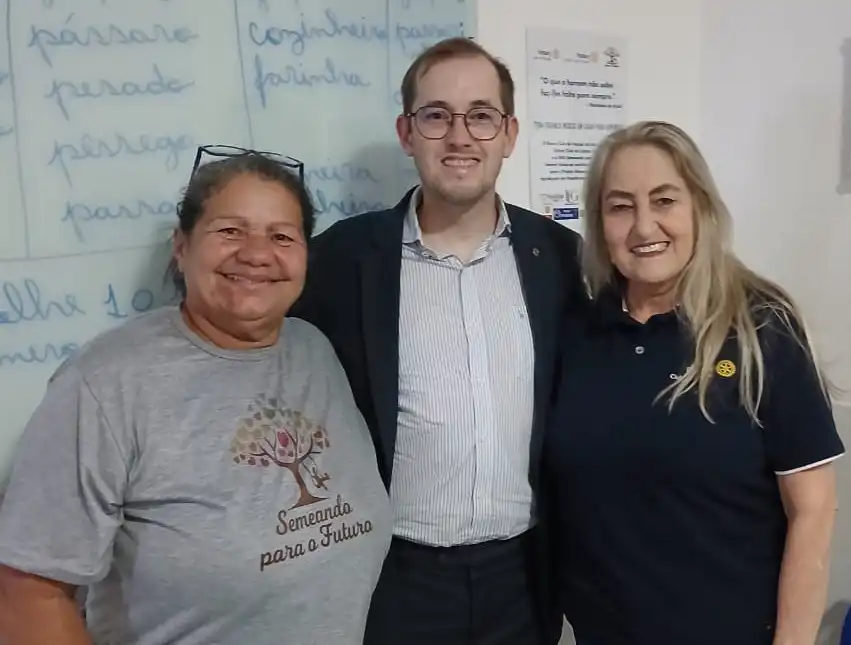 Três pessoas posam sorrindo dentro de uma sala de aula. Ao centro, o presidente do Rotary de Itaguaí, Josephj Piñeiro, de terno e óculos; à esquerda, Adeilda, presidente da ONG, que veste uma camiseta com a identificação da entidade “Semeando para o Futuro”; à direita, Eliane, mãe de Joseph e também rotariana, vestindo uma camisa polo com símbolo do Rotary. Ao fundo a lousa doada com palavras escritas, indicando ambiente educacional.