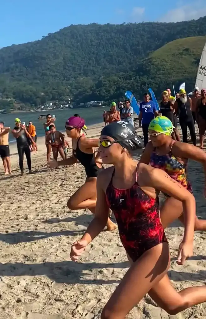 Nadadores usando toucas e roupas de competição correm pela areia em direção ao mar no início de uma prova de águas abertas. Ao fundo, há montanhas cobertas por vegetação, embarcações e espectadores acompanhando o evento na praia