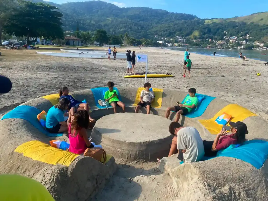 Grupo de jovens reunido em área de convivência construída na areia da Praia do Saco, em Mangaratiba, durante o 3º Festival Náutico, com montanhas, mar e embarcações esportivas ao fundo