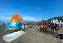 Estrutura do 3º Festival Náutico montada na Praia do Saco, em Mangaratiba, com participantes circulando pela areia, embarcações à vela ao fundo e painel da Vale em destaque durante evento esportivo realizado à beira-mar