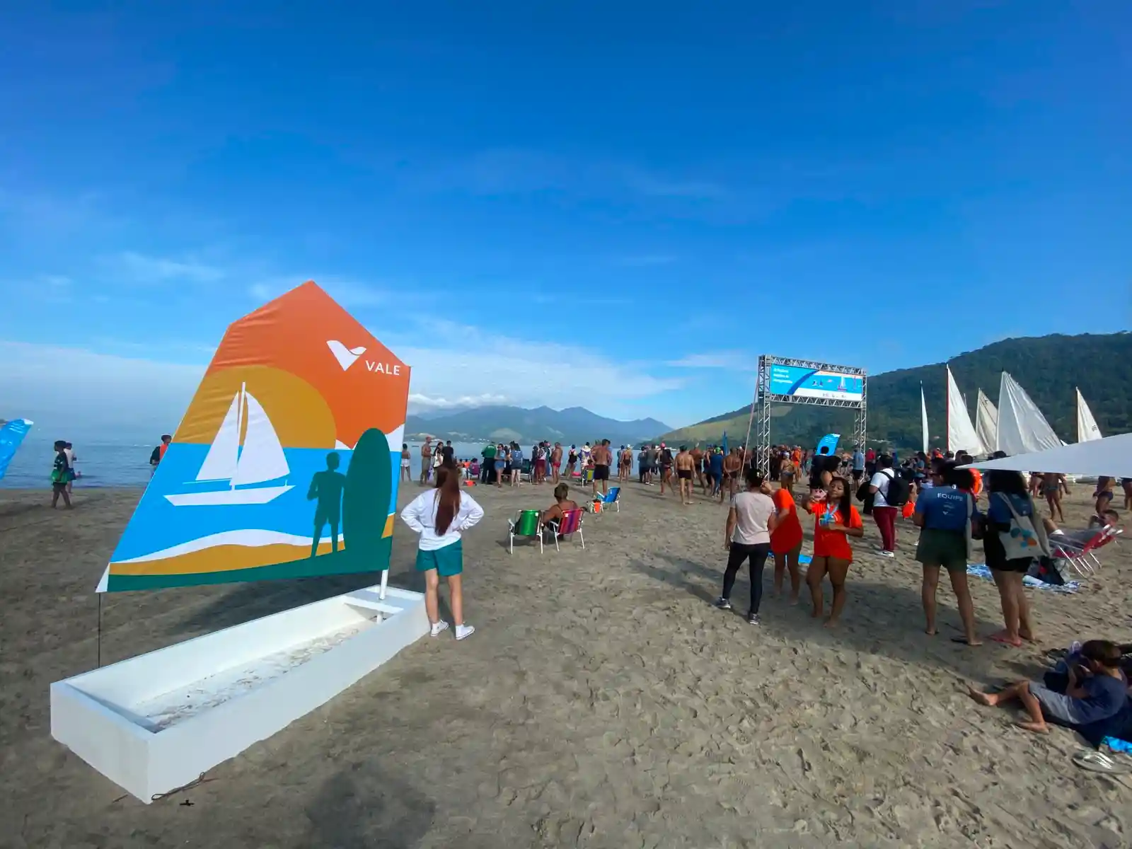 Estrutura do 3º Festival Náutico montada na Praia do Saco, em Mangaratiba, com participantes circulando pela areia, embarcações à vela ao fundo e painel da Vale em destaque durante evento esportivo realizado à beira-mar
