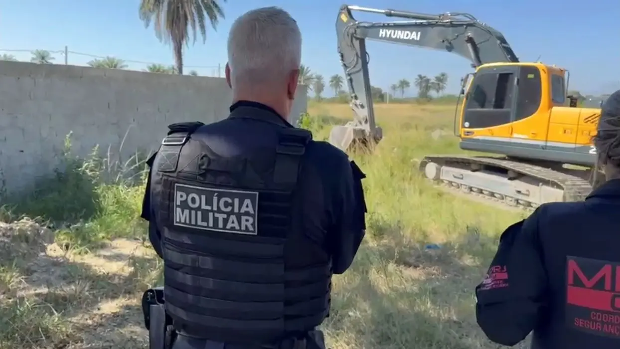 Agente da Polícia Militar observa uma retroescavadeira derrubando uma construção irregular