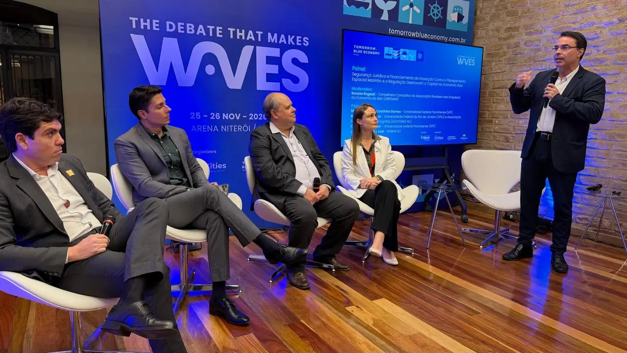 Participantes do painel “Segurança Jurídica e Capital Azul” debatem temas relacionados à Economia Azul durante o lançamento do Tomorrow.Blue Economy 2026, em Niterói. Quatro especialistas estão sentados no palco enquanto um palestrante fala ao público, diante de um painel com a identidade visual do evento.