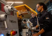 Estudante da Escola Técnica Roberto Rocca opera um torno mecânico durante atividade prática em laboratório, utilizando óculos de proteção e uniforme da instituição. Ao fundo, outros alunos acompanham o exercício. A imagem ilustra a formação técnica oferecida pela escola, mantida pela Ternium, com foco na capacitação de jovens para a indústria.
