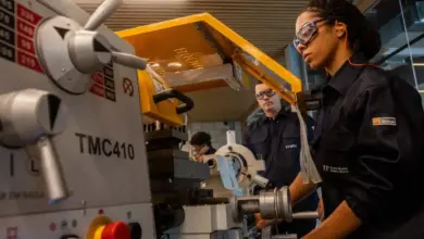 Estudante da Escola Técnica Roberto Rocca opera um torno mecânico durante atividade prática em laboratório, utilizando óculos de proteção e uniforme da instituição. Ao fundo, outros alunos acompanham o exercício. A imagem ilustra a formação técnica oferecida pela escola, mantida pela Ternium, com foco na capacitação de jovens para a indústria.
