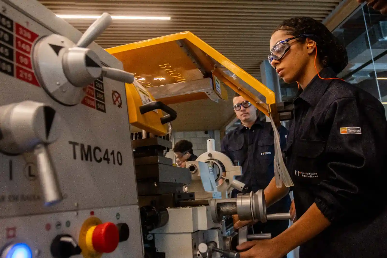 Estudante da Escola Técnica Roberto Rocca opera um torno mecânico durante atividade prática em laboratório, utilizando óculos de proteção e uniforme da instituição. Ao fundo, outros alunos acompanham o exercício. A imagem ilustra a formação técnica oferecida pela escola, mantida pela Ternium, com foco na capacitação de jovens para a indústria.