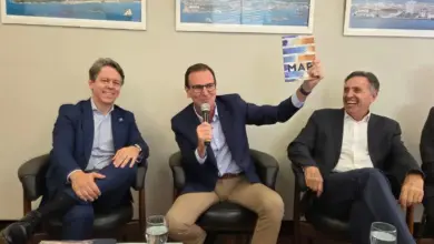 Facerj apresenta demanda ao Eduardo Paes, pré-candidato ao Governo do Estado do Rio de Janeiro, que aparece sentado ao centro da foto, falando ao microfone enquanto apresenta o livro “Economia do Mar”. Ao seu lado, representantes do setor empresarial acompanham o debate. O ambiente é decorado com fotografias de paisagens costeiras e portuárias, reforçando a temática do evento