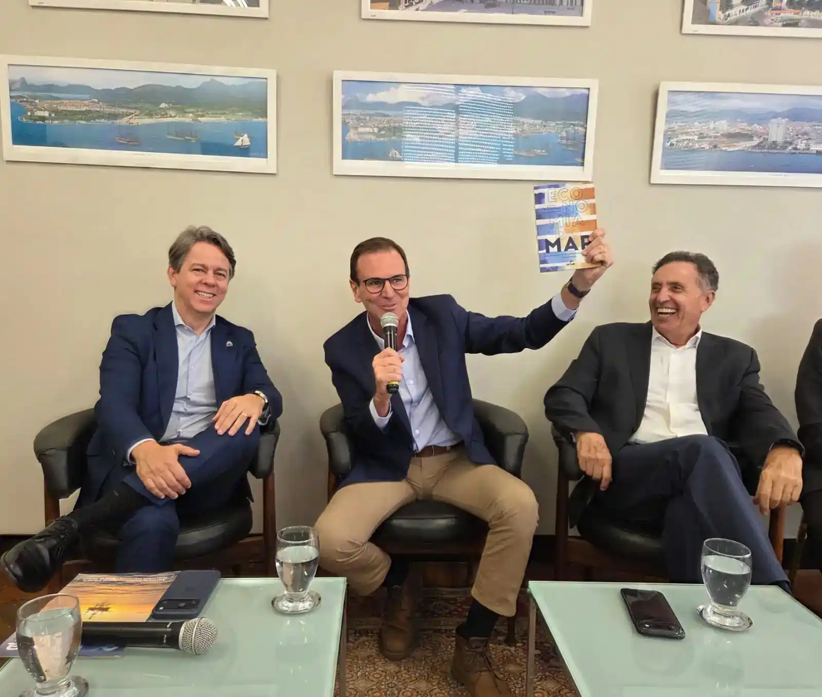 Facerj apresenta demanda ao Eduardo Paes, pré-candidato ao Governo do Estado do Rio de Janeiro, que aparece sentado ao centro da foto, falando ao microfone enquanto apresenta o livro “Economia do Mar”. Ao seu lado, representantes do setor empresarial acompanham o debate. O ambiente é decorado com fotografias de paisagens costeiras e portuárias, reforçando a temática do evento