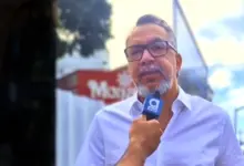 Marcio Canella está falando ao microfone identificado com a logo marca do jornal atual.