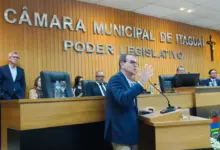 Eduardo Paes discursa de perfil no púlpito da Câmara Municipal de Itaguaí. Ele veste blazer azul-marinho, usa óculos e gesticula com a mão esquerda levantada. Ao fundo, uma parede de madeira exibe as letras prateadas "CÂMARA MUNICIPAL DE ITAGUAÍ - PODER LEGISLATIVO", com um crucifixo à direita. Atrás do político, vereadores e espectadores assistem sentados à solenidade.