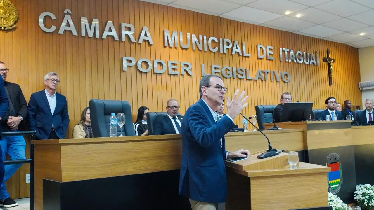 Eduardo Paes discursa de perfil no púlpito da Câmara Municipal de Itaguaí. Ele veste blazer azul-marinho, usa óculos e gesticula com a mão esquerda levantada. Ao fundo, uma parede de madeira exibe as letras prateadas "CÂMARA MUNICIPAL DE ITAGUAÍ - PODER LEGISLATIVO", com um crucifixo à direita. Atrás do político, vereadores e espectadores assistem sentados à solenidade.