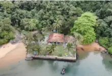 Vista aérea da Fazenda Marinha, no Terminal Ilha Guaíba (TIG), mostrando uma construção com telhado cerâmico cercada por área gramada, palmeiras e densa vegetação de Mata Atlântica. O espaço fica em uma pequena península às margens de águas calmas, com faixas de areia nas laterais e uma embarcação atracada em frente ao píer