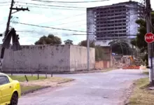 Poste de energia inclinado com telha de amianto presa à fiação elétrica em um cruzamento de Itaguaí após vendaval. Cabos estão caídos próximos à via, enquanto ao fundo há um prédio e uma máquina realizando serviços de derrubado de muro em uma rua interditada.