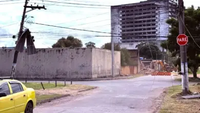 Poste de energia inclinado com telha de amianto presa à fiação elétrica em um cruzamento de Itaguaí após vendaval. Cabos estão caídos próximos à via, enquanto ao fundo há um prédio e uma máquina realizando serviços de derrubado de muro em uma rua interditada.
