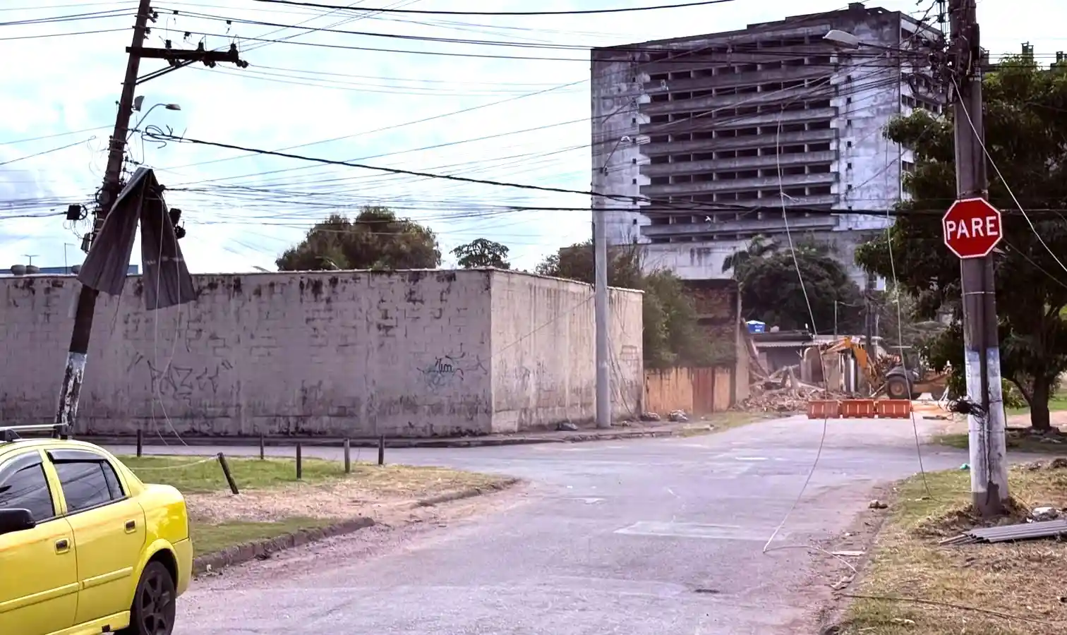 Poste de energia inclinado com telha de amianto presa à fiação elétrica em um cruzamento de Itaguaí após vendaval. Cabos estão caídos próximos à via, enquanto ao fundo há um prédio e uma máquina realizando serviços de derrubado de muro em uma rua interditada.