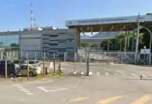 Claro, aqui está uma descrição do texto alt para a imagem: Uma fotografia ao nível da rua de um grande complexo industrial da ICN sob céu limpo. À esquerda, há um prédio administrativo moderno de dois andares com fachada de vidro e um padrão de painéis metálicos e de concreto em listras. Uma alta antena de comunicações de metal fica no telhado. Um pequeno estacionamento ao ar livre com vários carros está em frente, atrás de uma cerca de arame. No centro e à direita, um grande portão de entrada coberto para veículos tem um teto saliente apoiado por postes de metal. O letreiro no teto saliente lê: "Unidade de Fabricação de Estruturas Metálicas" e "800", com o logotipo da ICN visível. A entrada do portão é guardada por catracas e um portão de metal fechado com sinais de pare vermelhos. Além dos portões, há uma estrada asfaltada e mais infraestrutura industrial. O fundo apresenta montanhas arborizadas à distância. A estrada em primeiro plano tem marcas de faixa e símbolos de seta no asfalto.