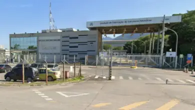 Claro, aqui está uma descrição do texto alt para a imagem: Uma fotografia ao nível da rua de um grande complexo industrial da ICN sob céu limpo. À esquerda, há um prédio administrativo moderno de dois andares com fachada de vidro e um padrão de painéis metálicos e de concreto em listras. Uma alta antena de comunicações de metal fica no telhado. Um pequeno estacionamento ao ar livre com vários carros está em frente, atrás de uma cerca de arame. No centro e à direita, um grande portão de entrada coberto para veículos tem um teto saliente apoiado por postes de metal. O letreiro no teto saliente lê: "Unidade de Fabricação de Estruturas Metálicas" e "800", com o logotipo da ICN visível. A entrada do portão é guardada por catracas e um portão de metal fechado com sinais de pare vermelhos. Além dos portões, há uma estrada asfaltada e mais infraestrutura industrial. O fundo apresenta montanhas arborizadas à distância. A estrada em primeiro plano tem marcas de faixa e símbolos de seta no asfalto.