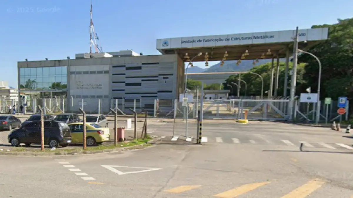 Claro, aqui está uma descrição do texto alt para a imagem: Uma fotografia ao nível da rua de um grande complexo industrial da ICN sob céu limpo. À esquerda, há um prédio administrativo moderno de dois andares com fachada de vidro e um padrão de painéis metálicos e de concreto em listras. Uma alta antena de comunicações de metal fica no telhado. Um pequeno estacionamento ao ar livre com vários carros está em frente, atrás de uma cerca de arame. No centro e à direita, um grande portão de entrada coberto para veículos tem um teto saliente apoiado por postes de metal. O letreiro no teto saliente lê: "Unidade de Fabricação de Estruturas Metálicas" e "800", com o logotipo da ICN visível. A entrada do portão é guardada por catracas e um portão de metal fechado com sinais de pare vermelhos. Além dos portões, há uma estrada asfaltada e mais infraestrutura industrial. O fundo apresenta montanhas arborizadas à distância. A estrada em primeiro plano tem marcas de faixa e símbolos de seta no asfalto.