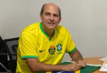 Alexandre Valle sentado à mesa, usando camisa amarela da Seleção Brasileira, em ambiente de escritório.