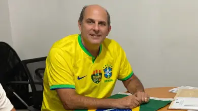 Alexandre Valle sentado à mesa, usando camisa amarela da Seleção Brasileira, em ambiente de escritório.
