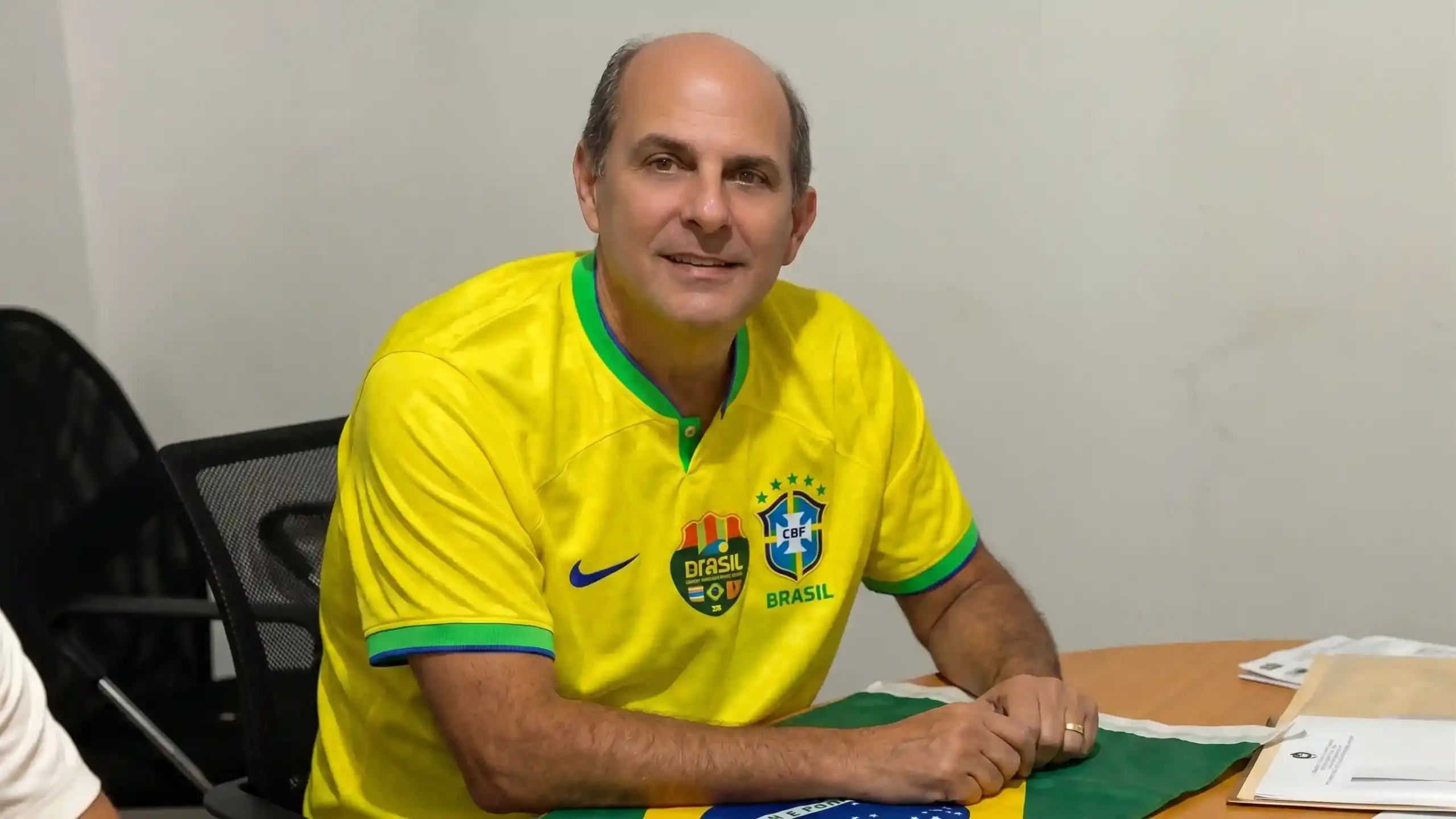 Alexandre Valle sentado à mesa, usando camisa amarela da Seleção Brasileira, em ambiente de escritório.