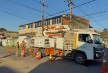 **Texto alternativo (alt):** Equipe da Light realiza manutenção na rede elétrica em um cruzamento de rua residencial, com caminhão equipado com cesto aéreo, dois eletricistas trabalhando próximo a um poste e área isolada por cones de sinalização. Ao fundo, há imóveis e fiação aérea.