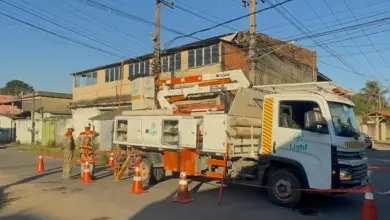 **Texto alternativo (alt):** Equipe da Light realiza manutenção na rede elétrica em um cruzamento de rua residencial, com caminhão equipado com cesto aéreo, dois eletricistas trabalhando próximo a um poste e área isolada por cones de sinalização. Ao fundo, há imóveis e fiação aérea.