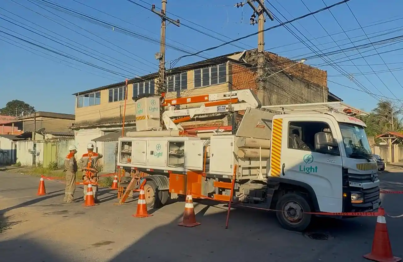 **Texto alternativo (alt):** Equipe da Light realiza manutenção na rede elétrica em um cruzamento de rua residencial, com caminhão equipado com cesto aéreo, dois eletricistas trabalhando próximo a um poste e área isolada por cones de sinalização. Ao fundo, há imóveis e fiação aérea.
