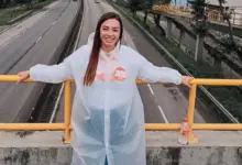 A imagem mostra Kelaine Gouulart de cabelos sorrindo e apoiada em um corrimão de metal amarelo em uma passarela sobre a rodovia Rio_santos. Ela usa uma capa de chuva branca transparente sobre uma camisa preta com um adesivo laranja "3080" no peito. A rodovia abaixo tem várias faixas de tráfego, com carros e caminhões em movimento. Ao fundo, árvores e prédios podem ser vistos. Kelaine está olhando para a câmera e parece estar desfrutando do dia. A imagem foi tirada de um ângulo ligeiramente elevado.