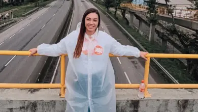 A imagem mostra Kelaine Gouulart de cabelos sorrindo e apoiada em um corrimão de metal amarelo em uma passarela sobre a rodovia Rio_santos. Ela usa uma capa de chuva branca transparente sobre uma camisa preta com um adesivo laranja "3080" no peito. A rodovia abaixo tem várias faixas de tráfego, com carros e caminhões em movimento. Ao fundo, árvores e prédios podem ser vistos. Kelaine está olhando para a câmera e parece estar desfrutando do dia. A imagem foi tirada de um ângulo ligeiramente elevado.