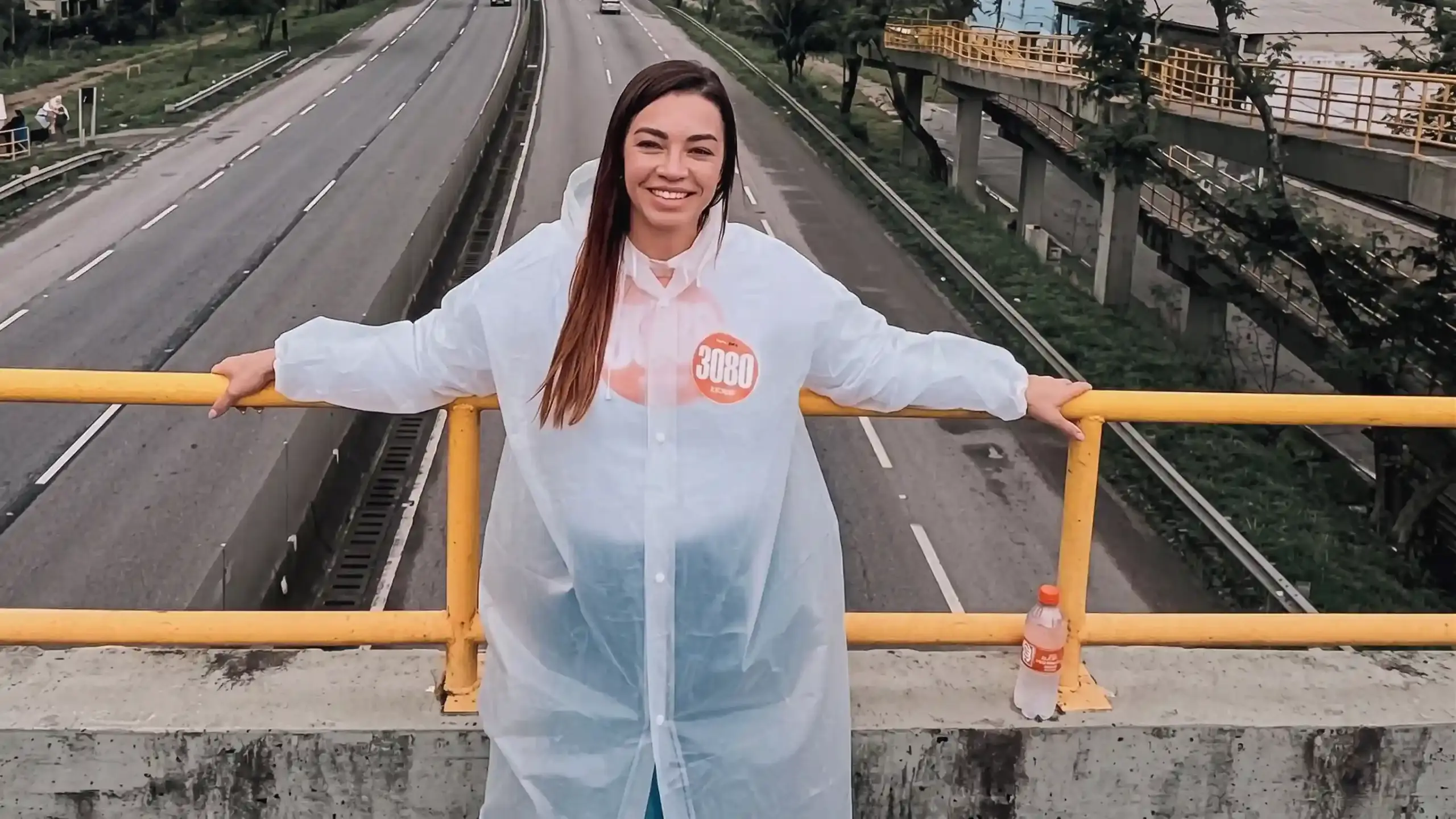 A imagem mostra Kelaine Gouulart de cabelos sorrindo e apoiada em um corrimão de metal amarelo em uma passarela sobre a rodovia Rio_santos. Ela usa uma capa de chuva branca transparente sobre uma camisa preta com um adesivo laranja "3080" no peito. A rodovia abaixo tem várias faixas de tráfego, com carros e caminhões em movimento. Ao fundo, árvores e prédios podem ser vistos. Kelaine está olhando para a câmera e parece estar desfrutando do dia. A imagem foi tirada de um ângulo ligeiramente elevado.