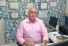 Dr. Helinho, homem de pele clara e calvo, veste camisa social rosa de mangas compridas e está sentado em uma mesa de escritório com papéis, um notebook e uma impressora preta. Ele olha em direção à câmera. Ao fundo, há uma parede com papel de parede estampado em tons de azul e branco, decorada com diversos quadros de diplomas e certificados emoldurados.
