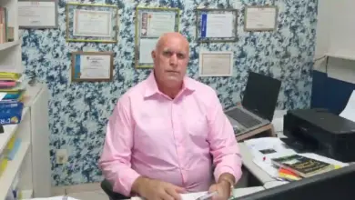 Dr. Helinho, homem de pele clara e calvo, veste camisa social rosa de mangas compridas e está sentado em uma mesa de escritório com papéis, um notebook e uma impressora preta. Ele olha em direção à câmera. Ao fundo, há uma parede com papel de parede estampado em tons de azul e branco, decorada com diversos quadros de diplomas e certificados emoldurados.