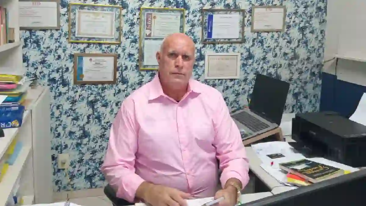 Dr. Helinho, homem de pele clara e calvo, veste camisa social rosa de mangas compridas e está sentado em uma mesa de escritório com papéis, um notebook e uma impressora preta. Ele olha em direção à câmera. Ao fundo, há uma parede com papel de parede estampado em tons de azul e branco, decorada com diversos quadros de diplomas e certificados emoldurados.