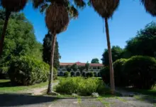 Prédio histórico da Universidade Rural com varanda em arcos, cercado por jardim, palmeiras e vegetação.