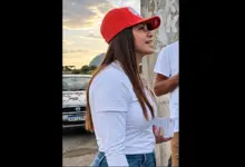Kelaine usando boné vermelho e camiseta branca, em pé ao lado de um muro; um carro aparece ao fundo e parte de outra pessoa está à direita. A foto está centralizada em duas colunas na cor preta.