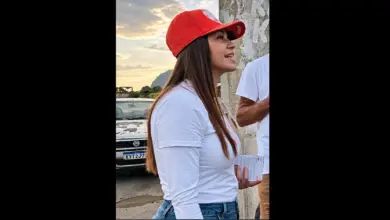 Kelaine usando boné vermelho e camiseta branca, em pé ao lado de um muro; um carro aparece ao fundo e parte de outra pessoa está à direita. A foto está centralizada em duas colunas na cor preta.