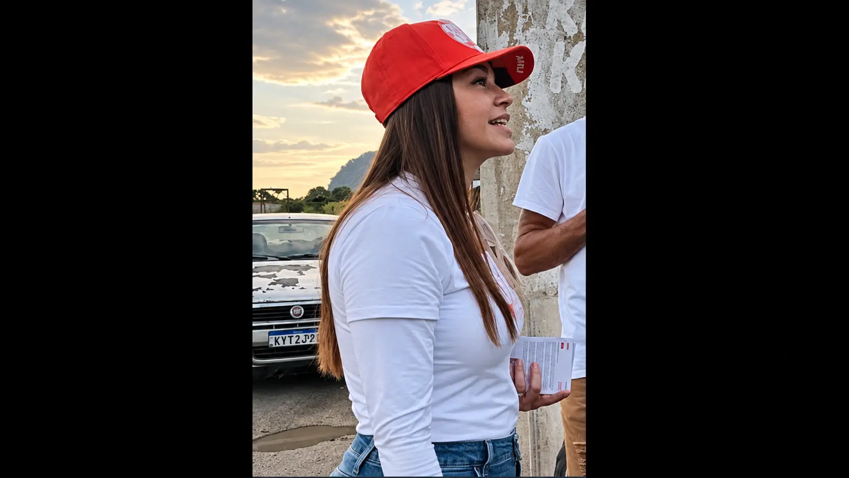 Kelaine usando boné vermelho e camiseta branca, em pé ao lado de um muro; um carro aparece ao fundo e parte de outra pessoa está à direita. A foto está centralizada em duas colunas na cor preta.