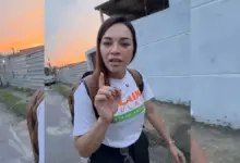 A candidata Kelaine Goulart caminha por uma rua de Itaguaí, olhando para a câmera e apontando o dedo indicador para cima enquanto fala. Ela veste uma camiseta branca de campanha e mochila nas costas, cercada por moradores e apoiadores ao ar livre no fim de tarde.